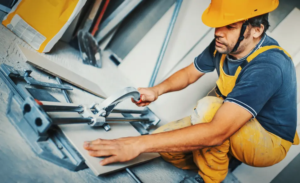 A professional renovator in a bright yellow safety vest and helmet using a heavy-duty tile cutter to split a tile into three parts, under an restricted area where flooring is being installed on the hard screed layer.