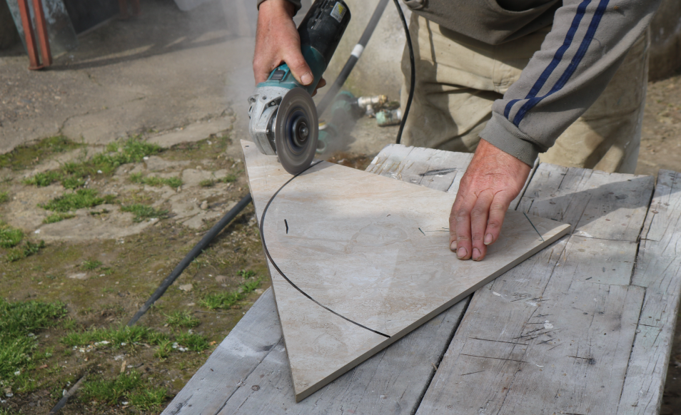 A professional flooring and wall installation contractor carefully guides the tile to be trimmed with an electrical round saw. As the tile itself is triangle in shape, a pencil marking has been drawn onto the tile as the contractor trims according to the inked surface. A rounded body is desired out of the current tile state.