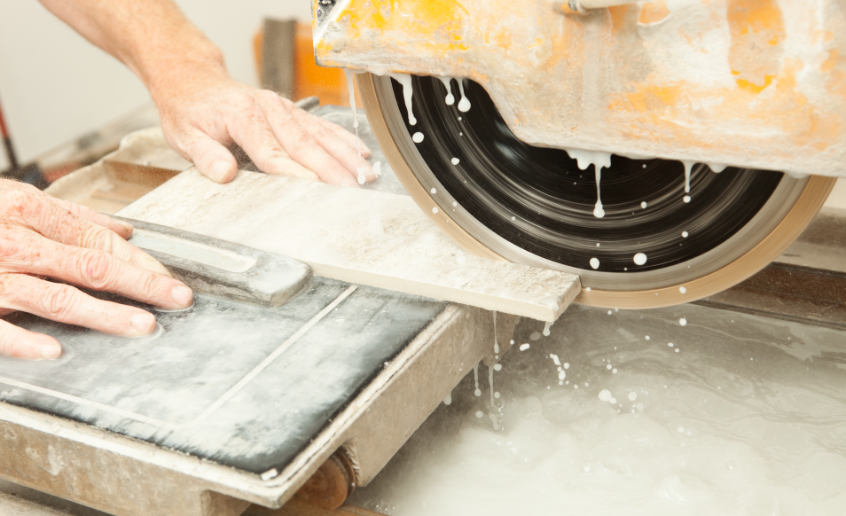 A smaller tile being placed and move into a circular saw by a pair of hands to be divided into two pieces. The entire process of both the tile and round saw is being drenched with water to facilitate better cutting precision while keeping the the saw blade cooled.